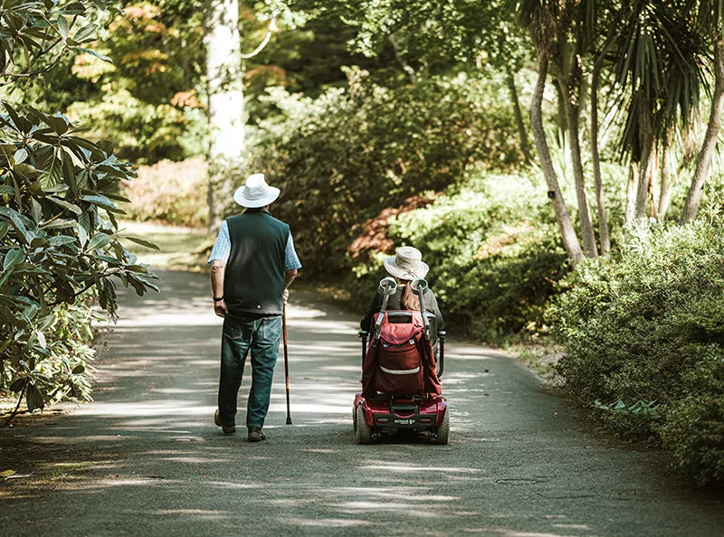 From the back we see a man with a cane and a woman with a mobility device on a path in a lush garden